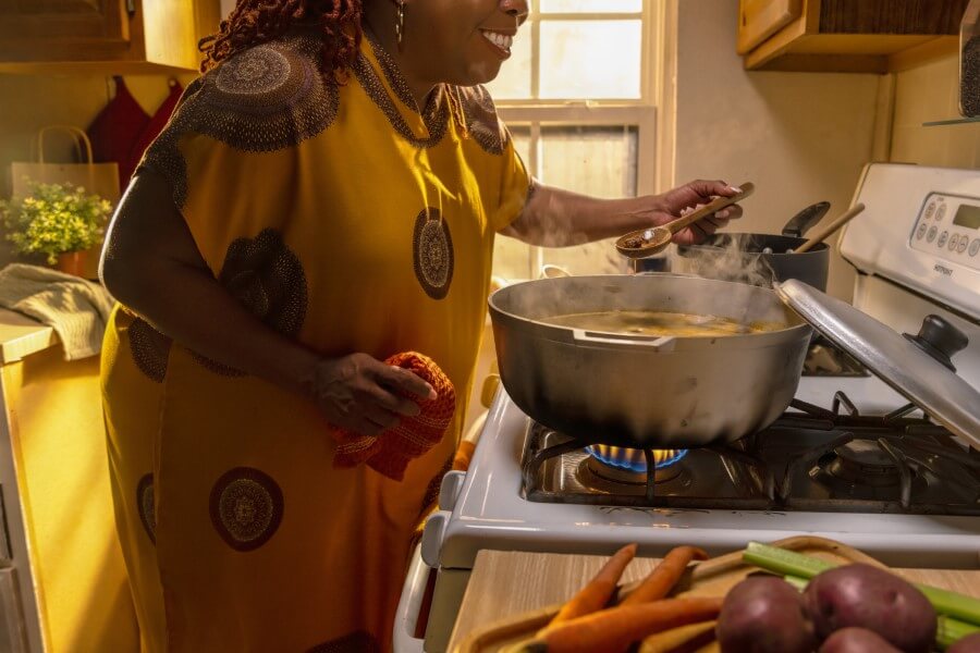 Black woman in a patterned yellow dress making stew in an apartement kitchen.